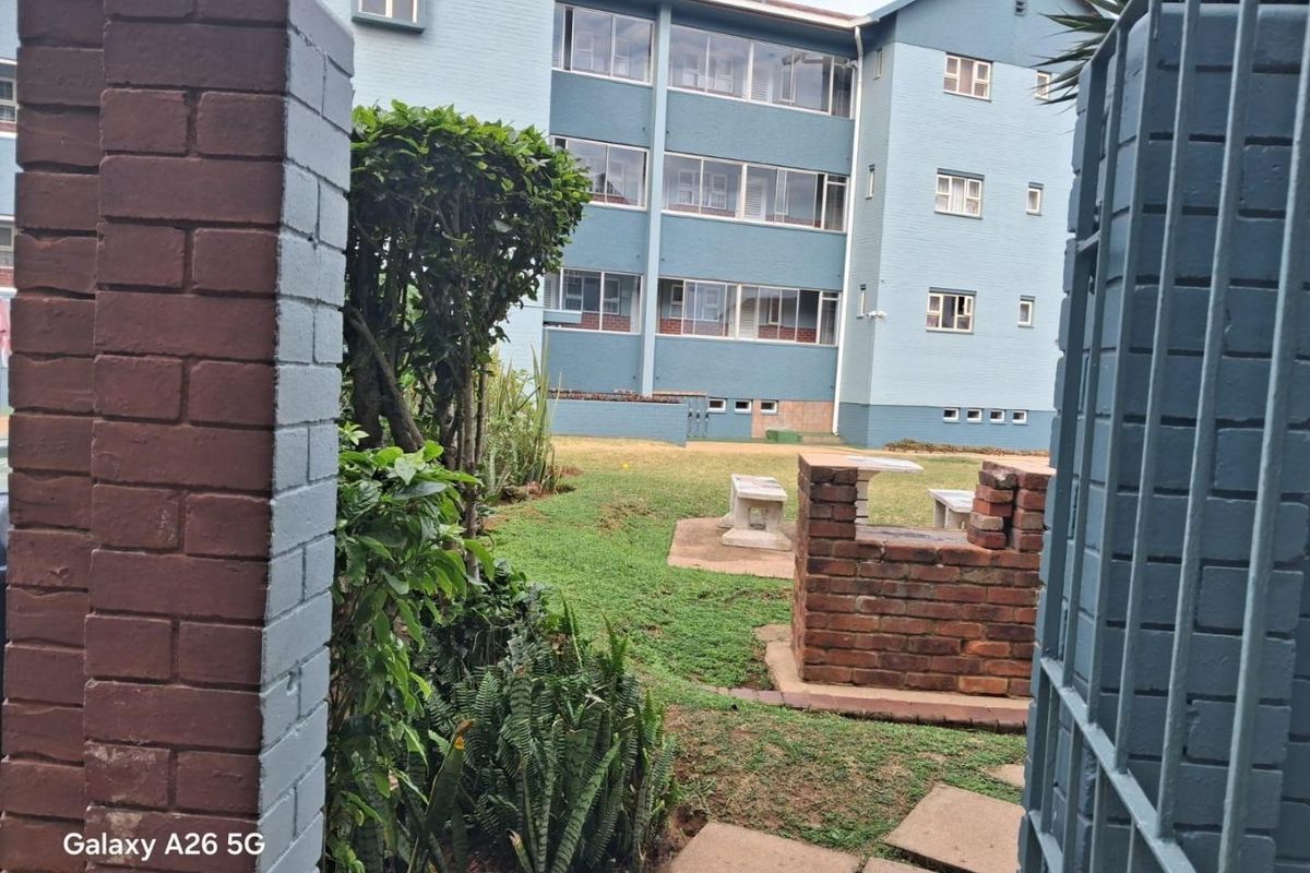View of a communal garden area featuring a brick braai, paved pathways, and a multi-story apartment building facade in the background.