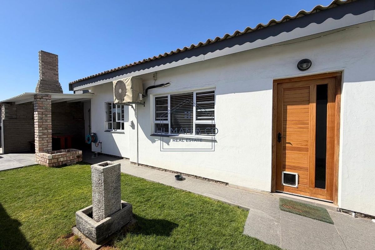 Rear exterior view featuring a paved patio area, built-in brick braai, lawn, and a wooden entrance door with a pet flap.