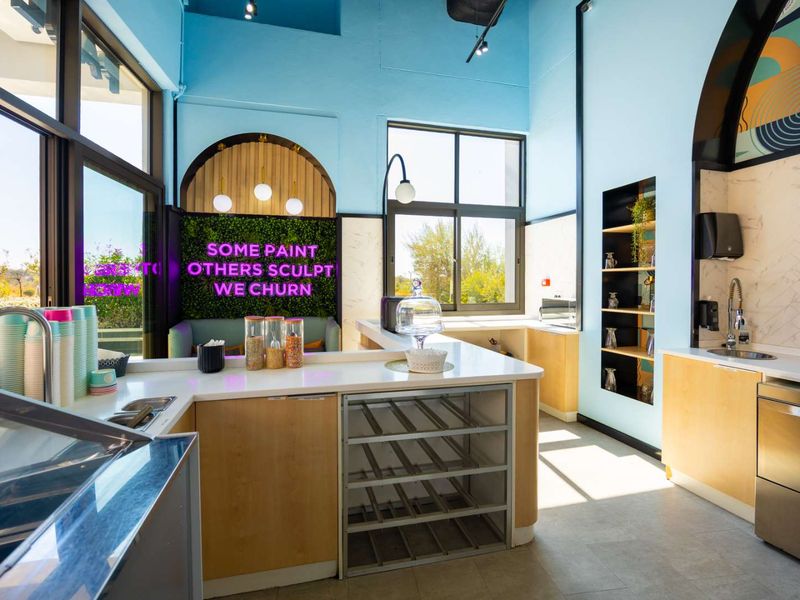 Modern commercial kitchen area featuring light wood cabinetry, stainless steel sink, integrated shelving, and large windows providing ample natural light.