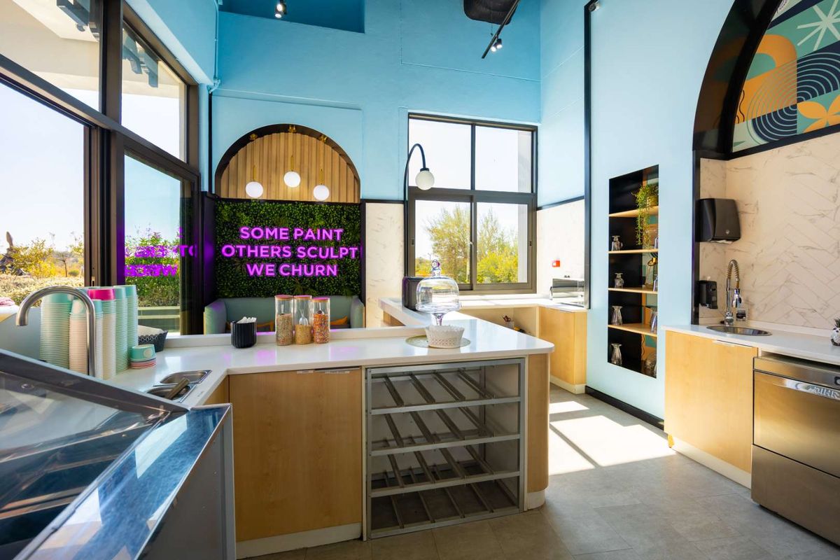 Modern commercial kitchen area featuring light wood cabinetry, stainless steel sink, integrated shelving, and large windows providing ample natural light.