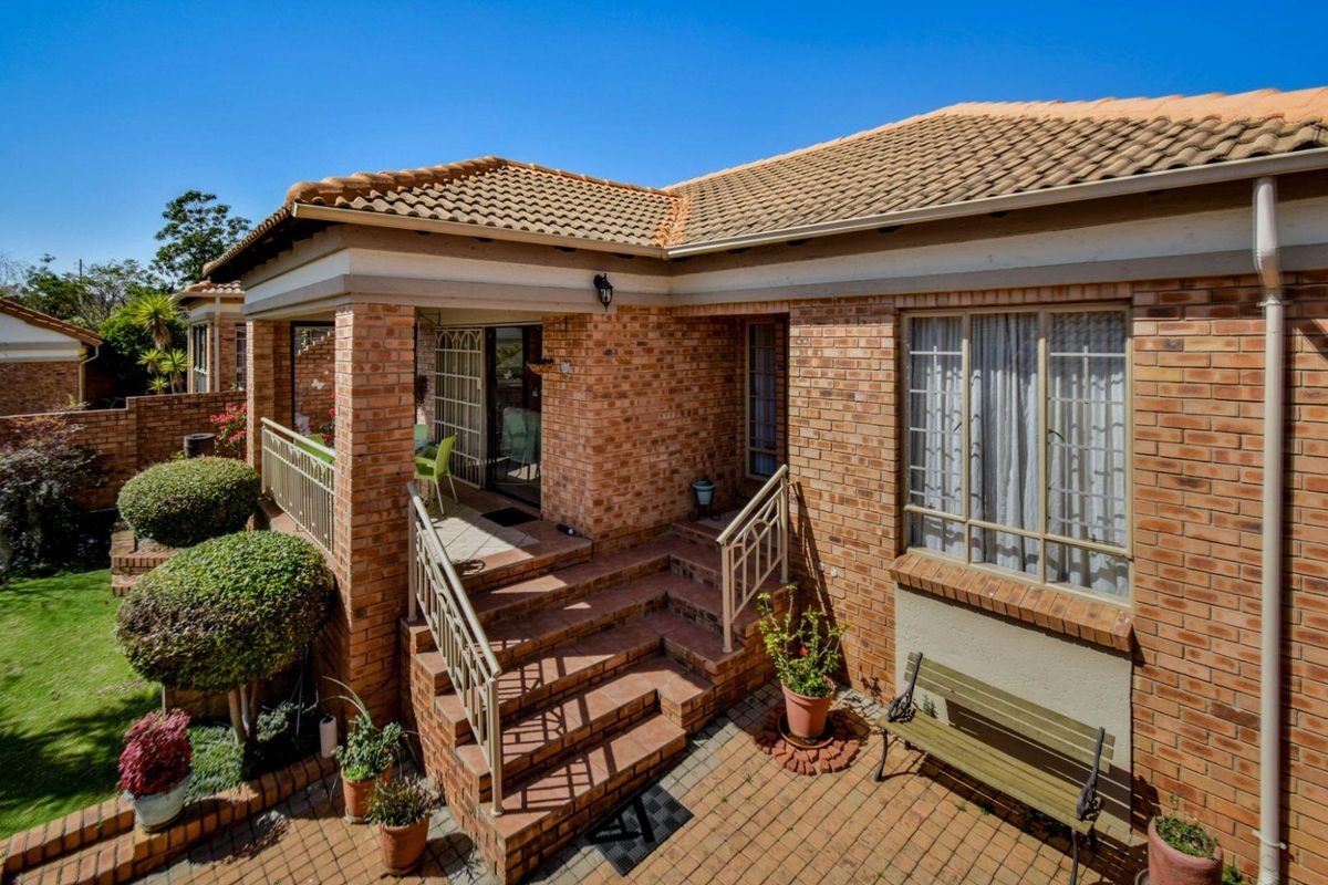 A well-maintained rear exterior view of a brick townhouse featuring a covered patio, paved courtyard area, and established garden surroundings.