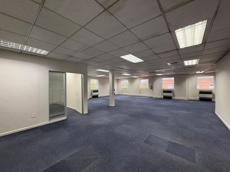 A spacious, open-plan office area featuring a suspended ceiling, recessed lighting, wall-mounted HVAC units, and blue carpet flooring.