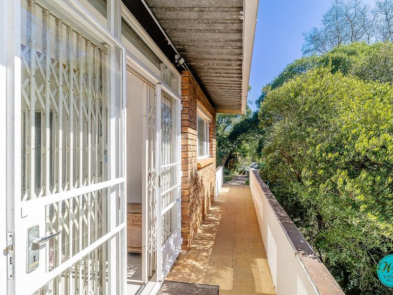A narrow exterior balcony featuring brick wall finishes, metal security gates, and a metal balustrade overlooking the surrounding garden area.