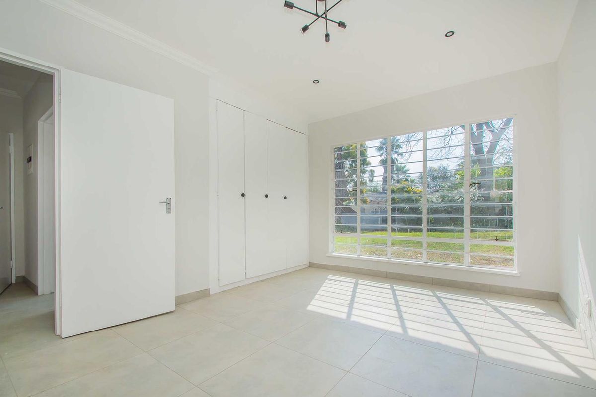 A bright, empty bedroom featuring large windows with security bars, built-in cupboards, and neutral tiled flooring.