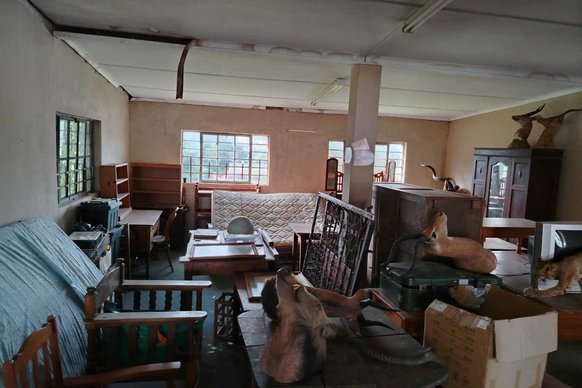 A cluttered interior space featuring large windows, exposed structural column, and dated ceiling finishes, requiring significant renovation and clearing to restore functionality.