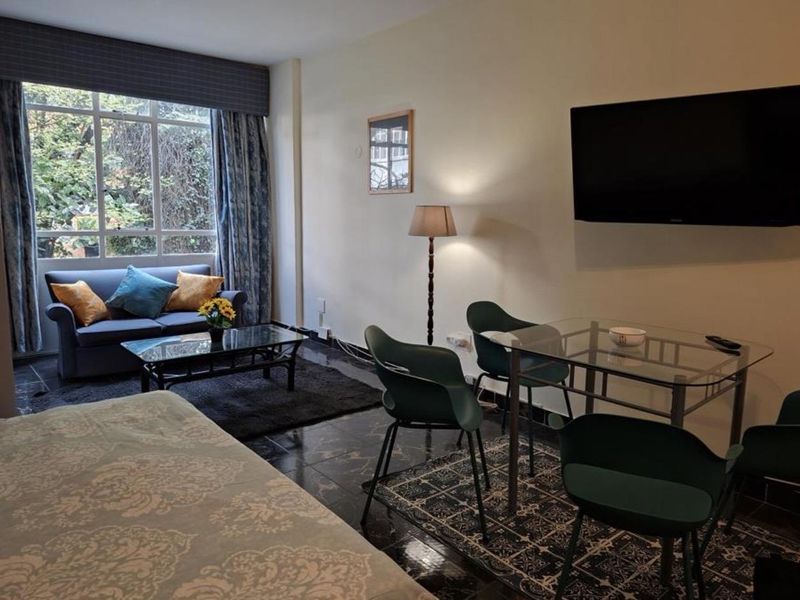 An open-plan living and dining area featuring large windows, dark tiled flooring, and a wall-mounted television, offering a functional and versatile living space.