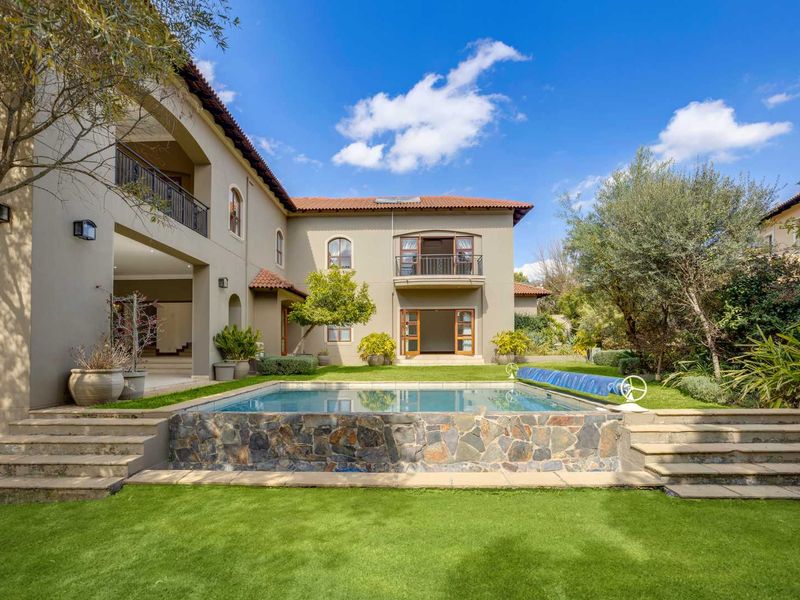 A well-maintained rear exterior view featuring a swimming pool with stone cladding, a landscaped garden, and a two-story house with a balcony and patio.