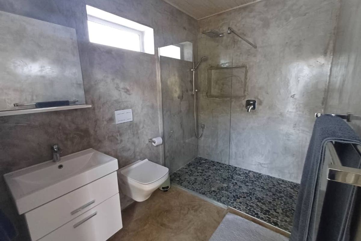 Modern bathroom featuring a walk-in shower with pebble flooring, wall-mounted toilet, vanity basin, and polished concrete-style wall finishes.