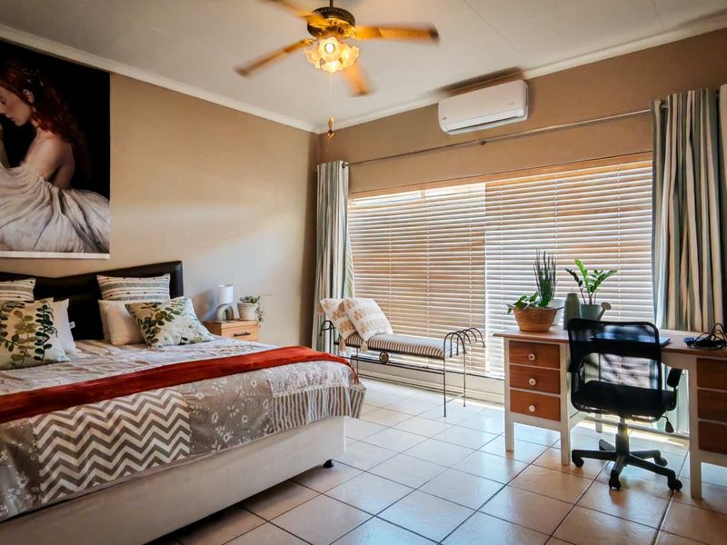 A spacious bedroom featuring tiled flooring, a ceiling fan, and a wall-mounted split-system air conditioner, with large windows fitted with venetian blinds.