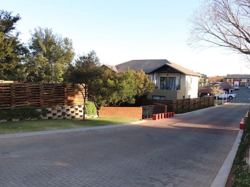 A street-level view of the townhouse complex entrance, featuring paved driveways, landscaped garden areas, and well-maintained boundary fencing.