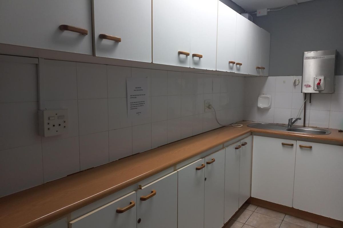 A functional office kitchenette featuring white cabinetry, laminate countertops, tiled splashbacks, a stainless steel sink, and a wall-mounted water boiler.