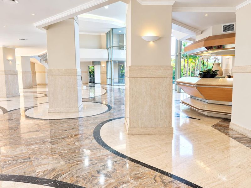 Spacious apartment building lobby featuring polished travertine flooring with decorative marble inlay, structural columns, and a built-in reception desk.