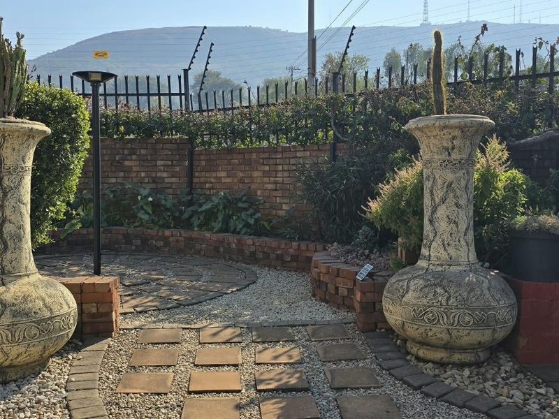A landscaped garden area featuring a paved walkway, gravel beds, brick retaining walls, and secure perimeter fencing with electric security wires.