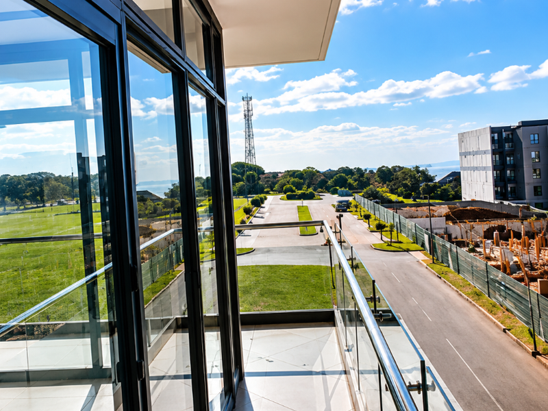 A modern, spacious balcony featuring a sleek glass balustrade, tiled flooring, and large glass sliding doors with views over the surrounding area.