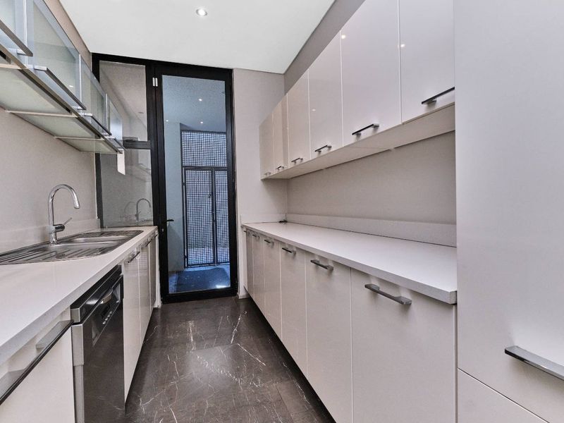 A modern, well-maintained scullery featuring sleek white cabinetry, stone countertops, a stainless steel sink, and integrated dishwasher with direct access to an outdoor area.