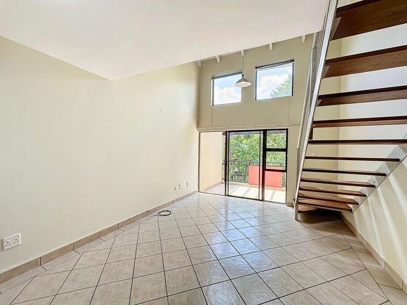 Spacious open-plan living area featuring double volume ceilings, tiled flooring, a wooden staircase, and direct access to a private balcony.