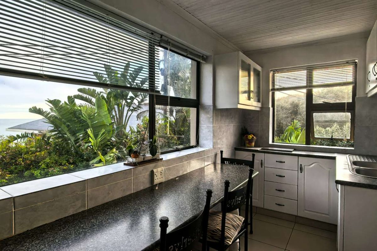A functional kitchen featuring built-in cabinetry, laminate countertops, and a double stainless steel sink, with large windows offering scenic garden and ocean views.