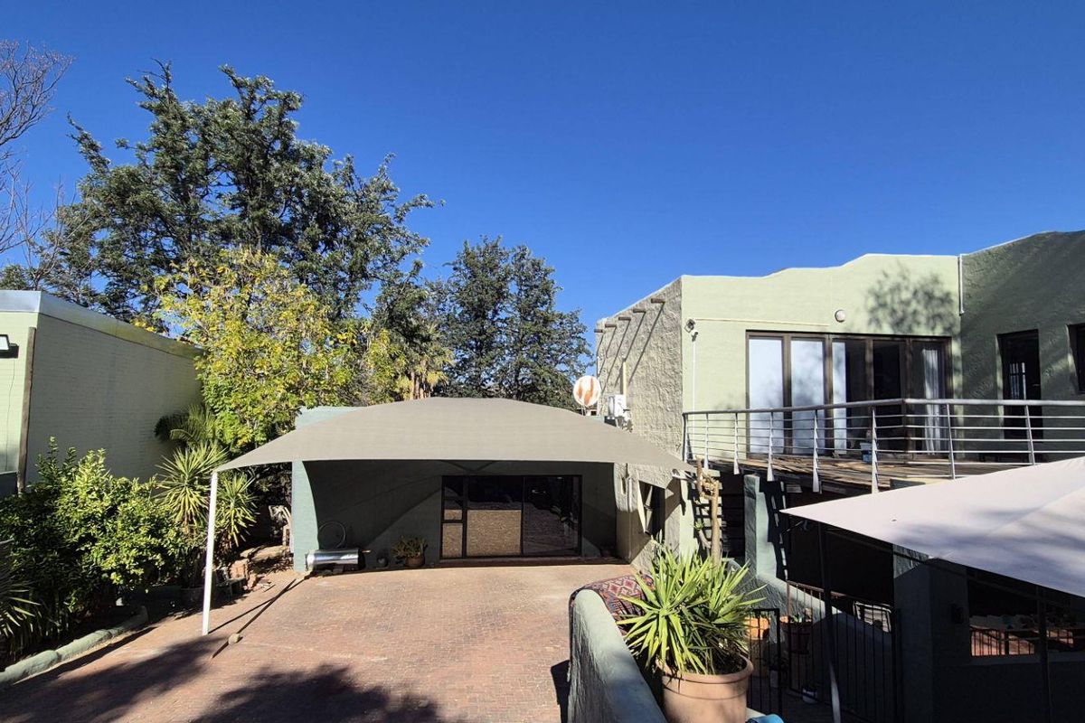 Rear exterior view of the property featuring a paved driveway with a shade sail, a balcony with metal railings, and sliding glass door access.