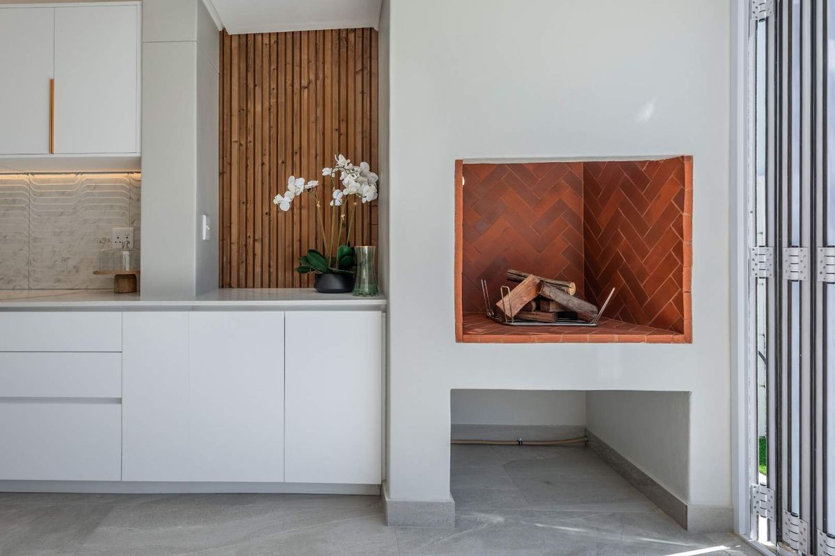 Modern indoor braai area featuring a built-in fireplace with herringbone brickwork, sleek white cabinetry, and timber slat wall paneling.