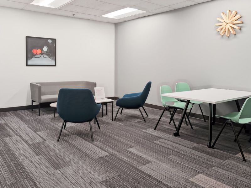A modern office breakout area featuring neutral walls, grey carpet tiles, and a suspended ceiling with recessed lighting.