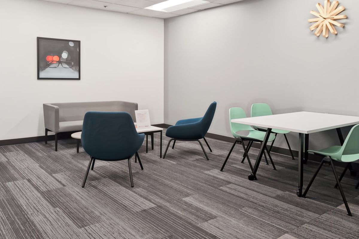 A modern office breakout area featuring neutral walls, grey carpet tiles, and a suspended ceiling with recessed lighting.