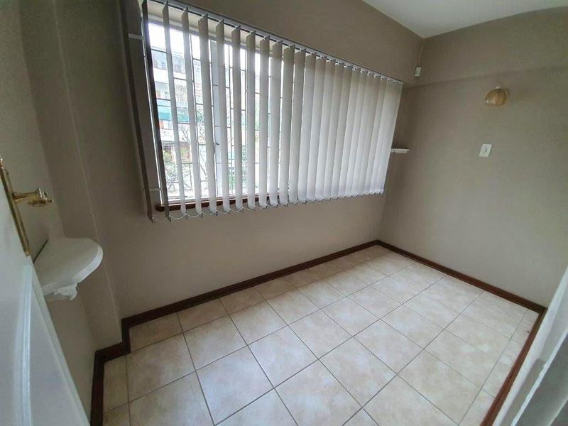 A bright, empty bedroom featuring neutral tiled flooring, vertical window blinds, and a wall-mounted light fixture, offering a clean canvas for personalization.