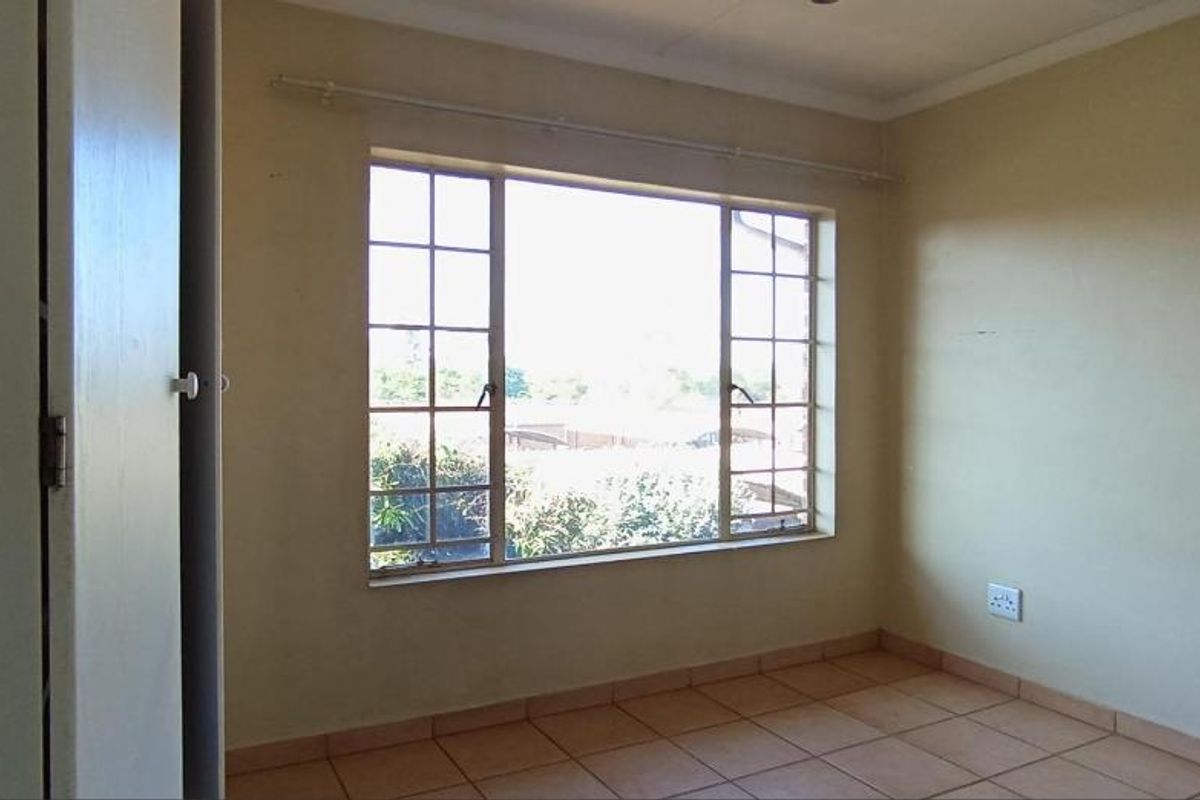 A bright, empty bedroom featuring tiled flooring, a large window with metal frames, and built-in cupboard storage.