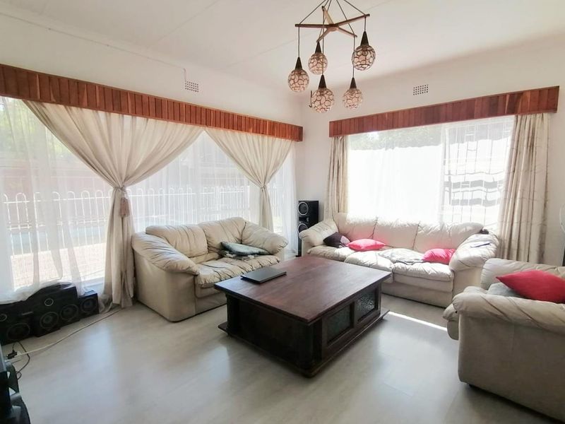 A bright living room featuring tiled flooring, large windows with wooden pelmets, and a decorative multi-pendant light fixture.