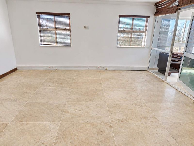 A bright, empty office space featuring neutral tiled flooring, large windows with venetian blinds, and direct access to an adjoining balcony area.