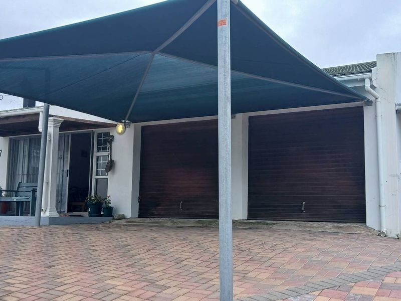 Front exterior view featuring a paved driveway, double garage with wooden doors, and a large shade sail covering the entrance area.