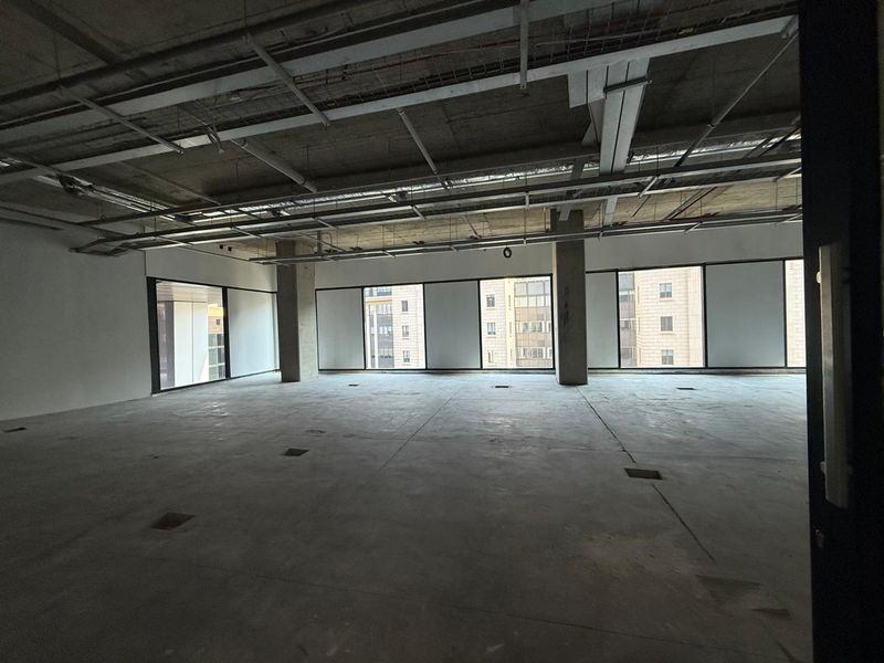 An open-plan office space in shell condition featuring exposed concrete ceilings, ducting, and large windows providing ample natural light.