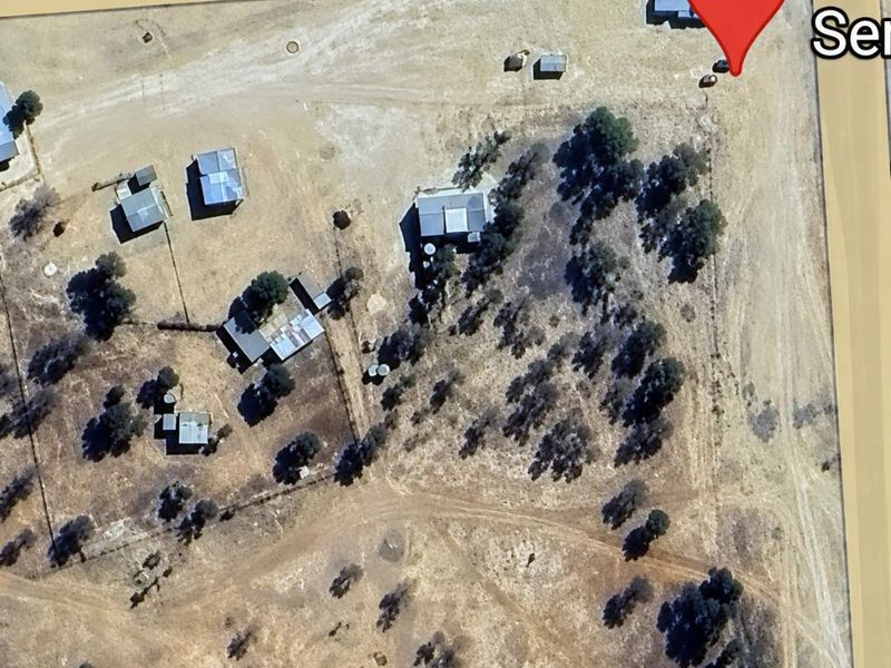 Aerial view of a residential land plot featuring multiple outbuildings, water tanks, and established trees with direct road access.