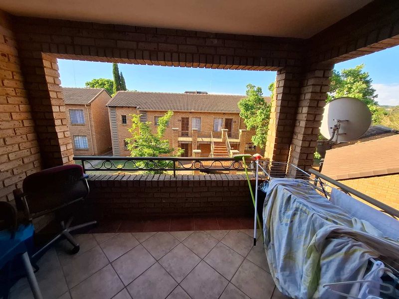 A covered balcony featuring brick pillars, tiled flooring, and a metal railing, overlooking the townhouse complex grounds.