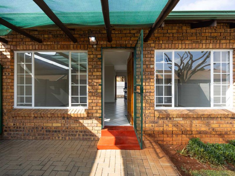 A paved patio area featuring a brick facade, shade cloth pergola, and a security gate leading into the interior of the townhouse.