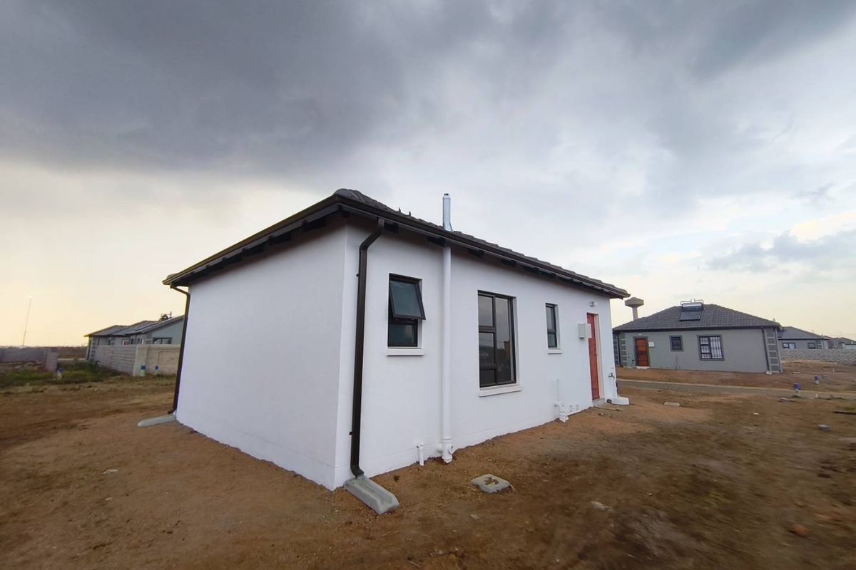 Exterior view of a modern single-story house featuring white rendered walls, dark window frames, and a tiled roof, set on an undeveloped plot.