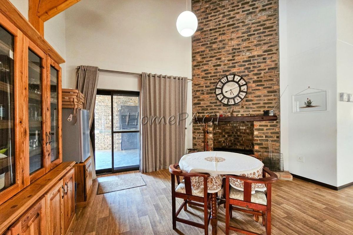 Spacious living area featuring a double-volume ceiling, exposed timber beams, and a prominent floor-to-ceiling face-brick fireplace with a wooden mantel.