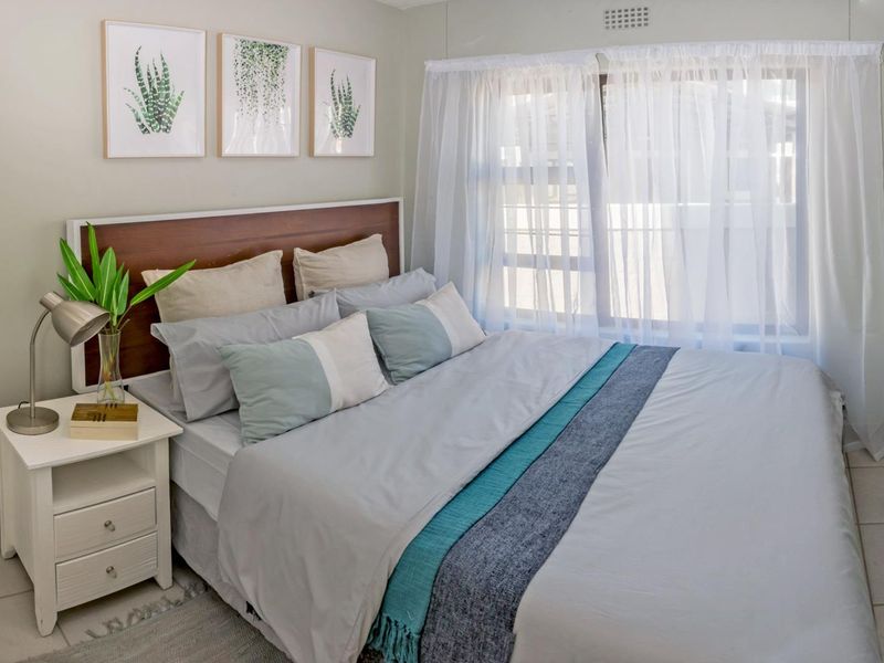 A well-lit bedroom featuring a built-in headboard, tiled flooring, and a large window with sheer curtains, providing a comfortable and functional living space.