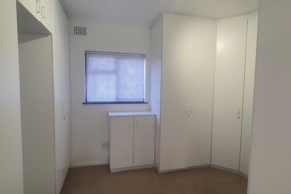 A clean, empty bedroom featuring extensive white built-in cabinetry, neutral carpeting, and a window with fitted blinds.