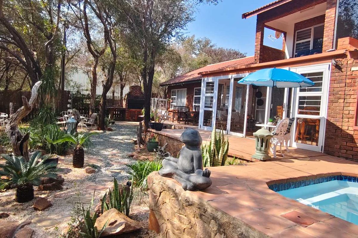 Rear exterior view featuring a brick house, tiled patio, swimming pool, and a landscaped garden with a built-in braai area.