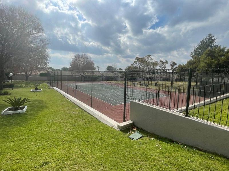 A well-maintained outdoor tennis court facility enclosed by perimeter fencing, set within a landscaped garden area.