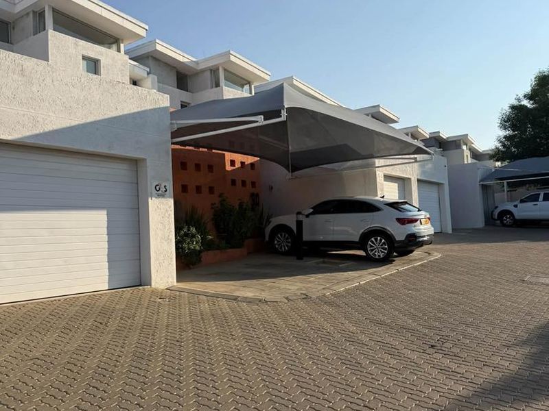 Front exterior view of a townhouse featuring a paved driveway, garage access, and a modern shade sail structure over the parking area.