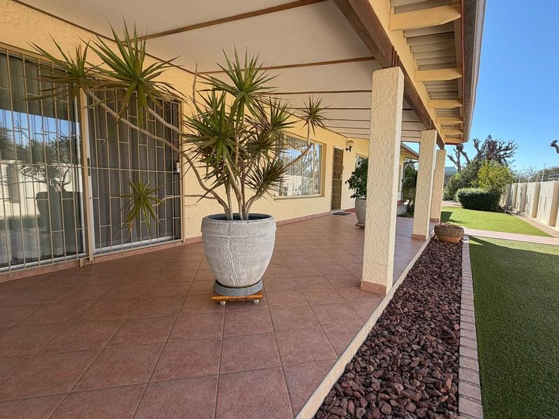 A spacious covered patio featuring tiled flooring, support pillars, and sliding glass doors, overlooking a well-maintained garden area.