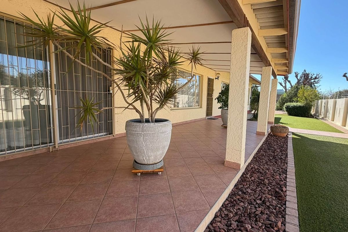 A spacious covered patio featuring tiled flooring, support pillars, and sliding glass doors, overlooking a well-maintained garden area.