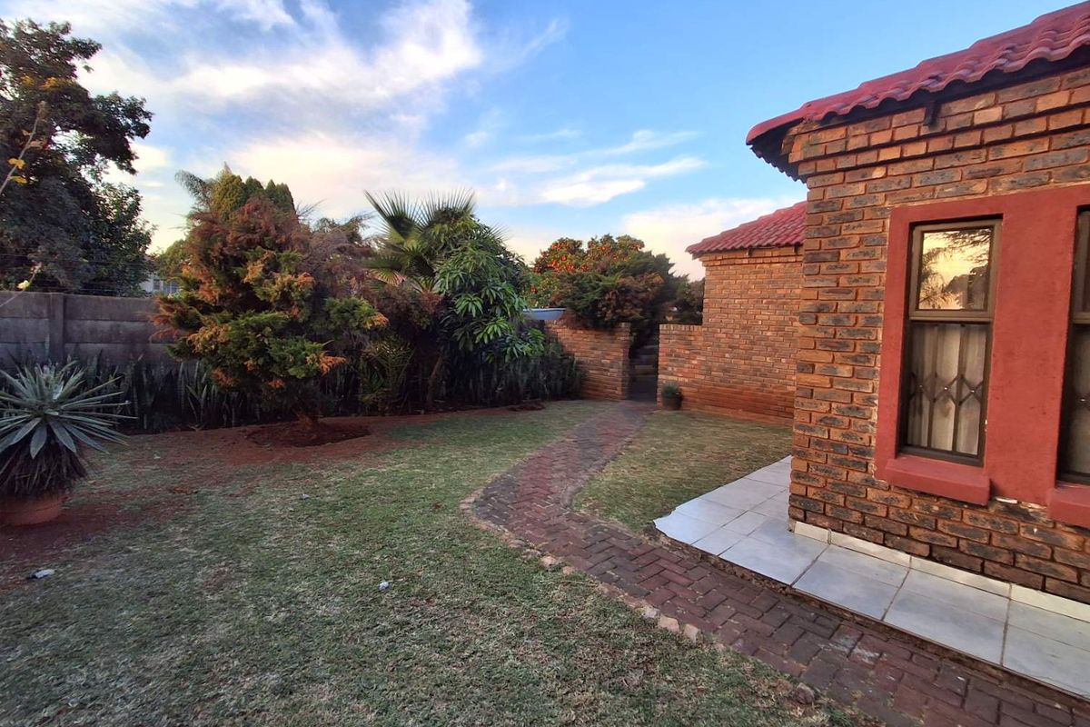 A view of the rear garden featuring a brick-paved pathway, established landscaping, and the exterior brick facade of the house.