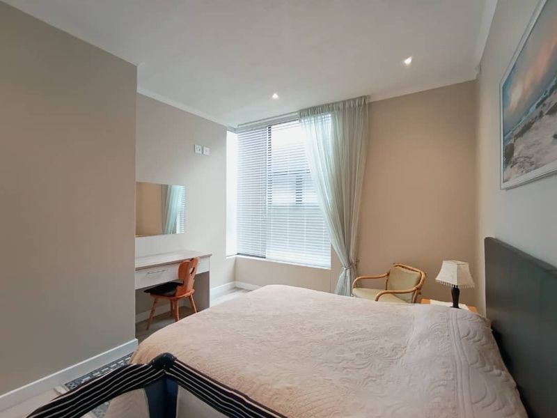 A well-maintained bedroom featuring neutral wall tones, recessed downlighting, and a built-in vanity area with large windows and venetian blinds.