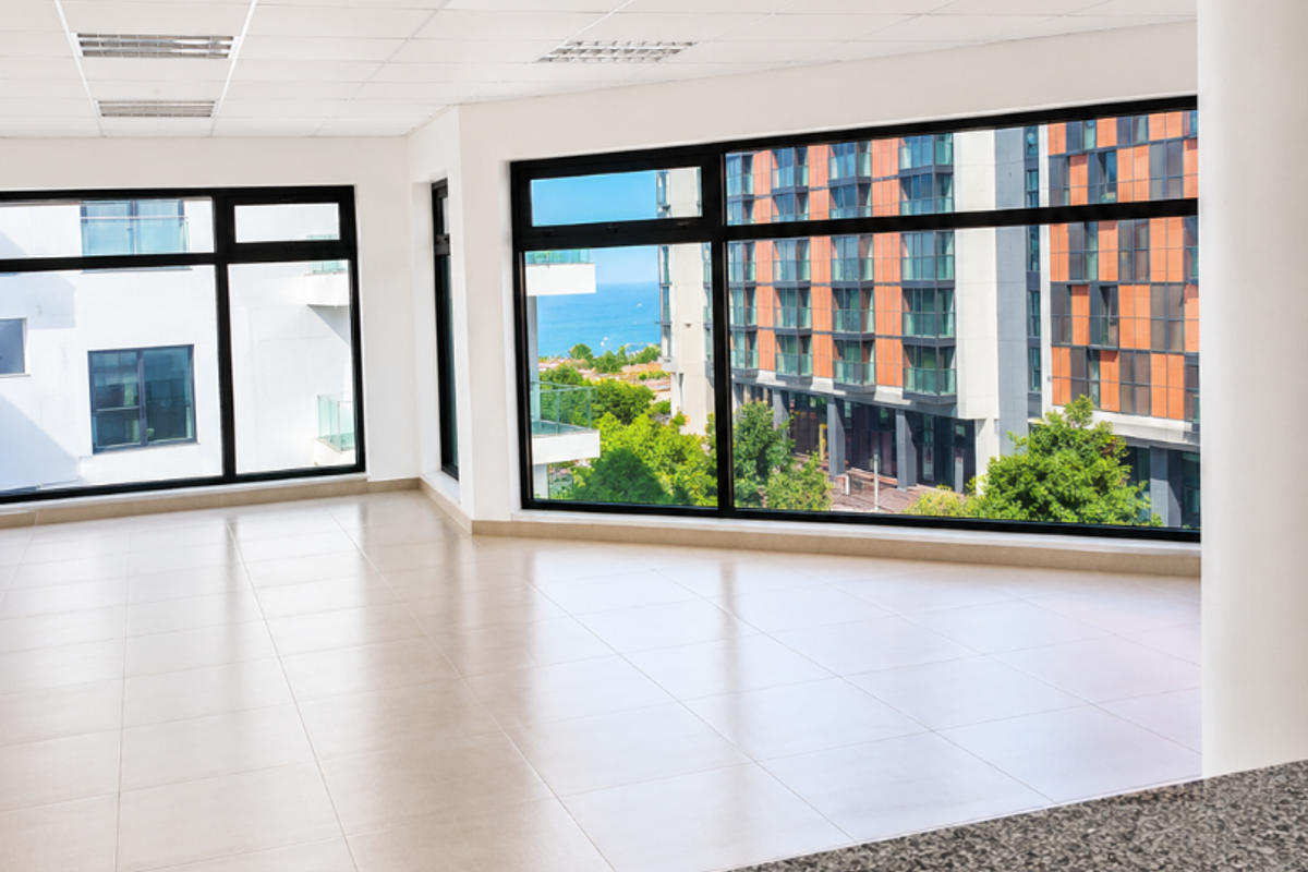 A bright, vacant office space featuring large windows with urban views, a suspended ceiling with recessed lighting, and a granite-topped service counter.