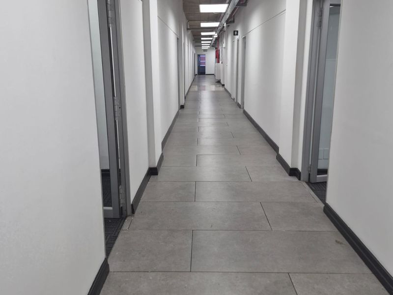 A clean, modern office hallway featuring tiled flooring, exposed concrete ceilings with integrated lighting, and exposed industrial-style cable trays.