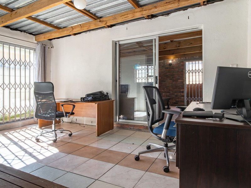 A functional office space featuring tiled flooring, exposed timber rafters, and a sliding glass door with security gate access.