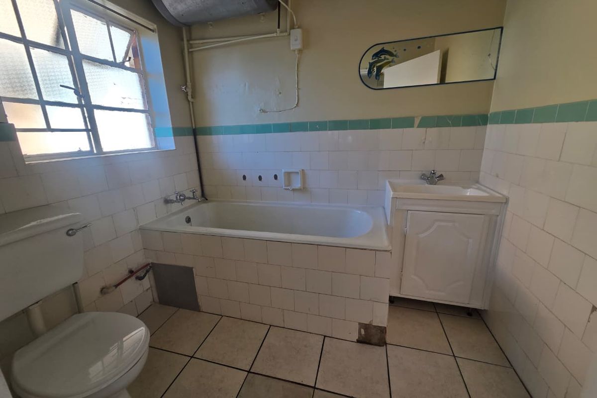 A dated bathroom featuring a standard bathtub, vanity basin, and toilet with original wall tiling and exposed water heater plumbing.