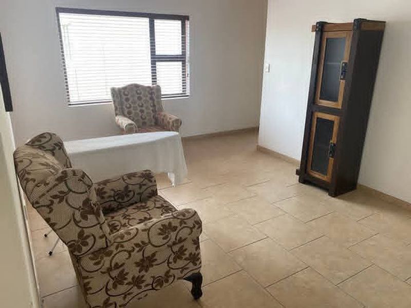 A living area featuring neutral tiled flooring and a large window with venetian blinds, offering a clean and functional space.